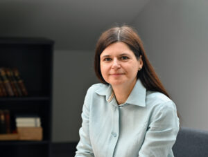 Besišypsanti tamsiaplaukė moteris, LAB direktorė Inga Davidonienė, vilkinti šviesiai melsvus marškinius. Ji sėdi patalpoje už stalo, fone matosi tamsios knygų lentynos.