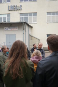 Ekskursijos dalyviai matomi iš nugaros, klausantys gido Alberto Kazlausko. Fone – didelis šviesus „Liregus“ gamybos pastatas su užrašu „Elektros instaliacija. Gamyba ir prekyba“.