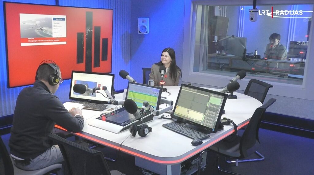 LRT radijo studijos nuotrauka: prie stalo sėdintis vedėjas kalbina besišypsančią viešnią, LAB direktorę, o fone matomas didelis raudonas ekranas ir langas į garso operatoriaus patalpą.