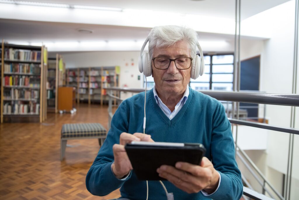 Bibliotekos erdvėje sėdintis žilplaukis vyras su didelėmis baltomis ausinėmis susikaupęs žiūri į planšetę.