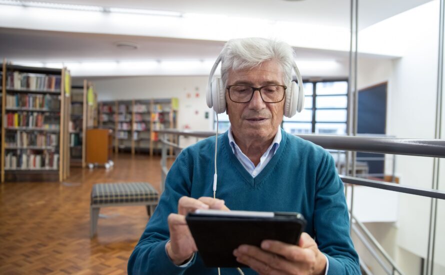 Bibliotekos erdvėje sėdintis žilplaukis vyras su didelėmis baltomis ausinėmis susikaupęs žiūri į planšetę.