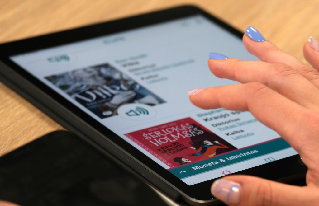 Stambiu planu pavaizduotas ant medinio stalo gulintis planšetinis kompiuteris. Moters ranka su šviesiai mėlynu manikiūru liečia ekraną, kuriame atverta Lietuvos audiosensorinės bibliotekos garsinių ir elektroninių knygų platforma ELVIS. Ekrane matomos knygų kortelės, tarp jų – raudonas knygos „Šerlokas Holmsas“ viršelis.