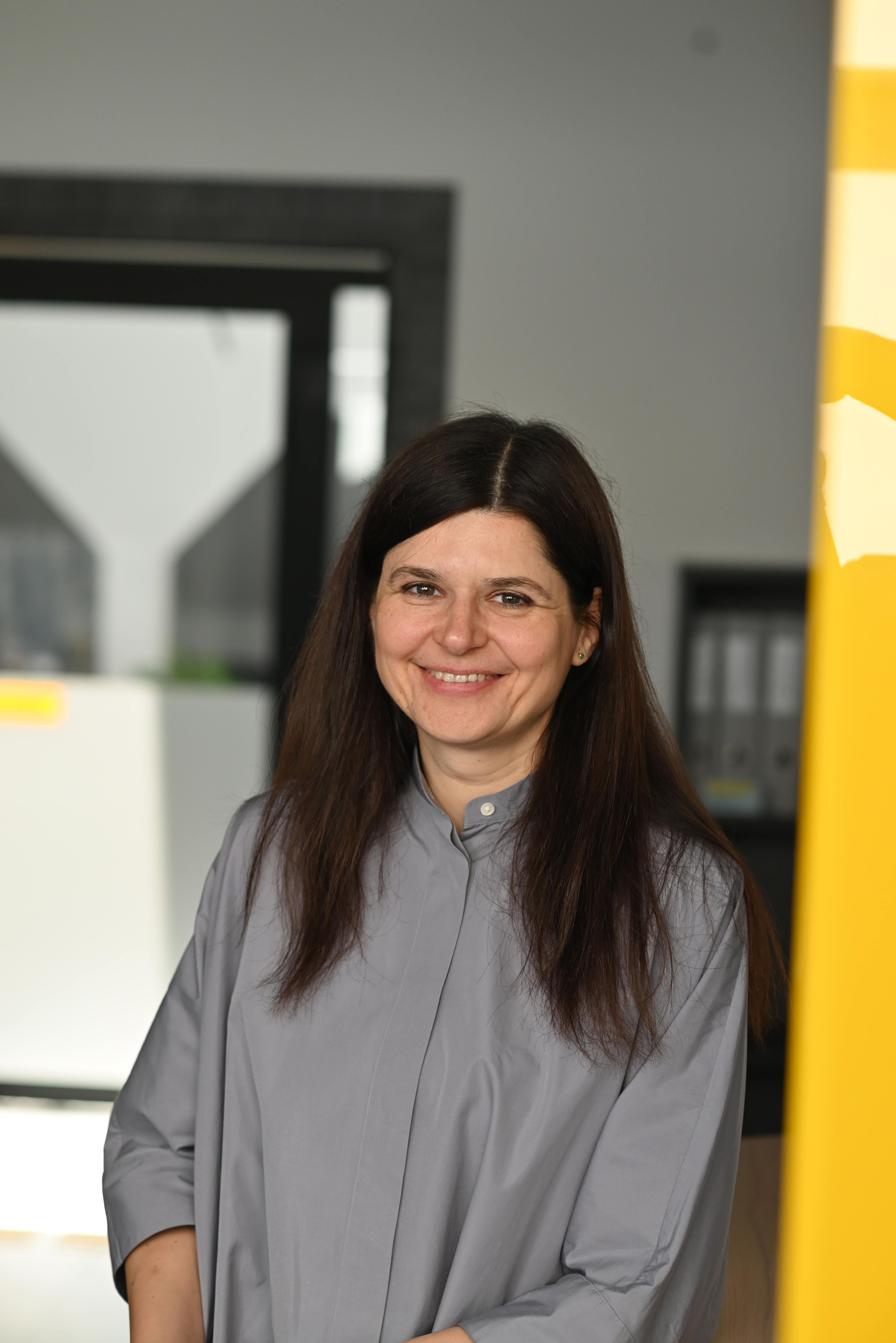 A portrait of Inga Davidonienė. A woman set against a bright office background, wearing a gray shirt and smiling broadly while looking directly into the camera. Her long, dark brown hair is parted down the middle and falls naturally over her shoulders.