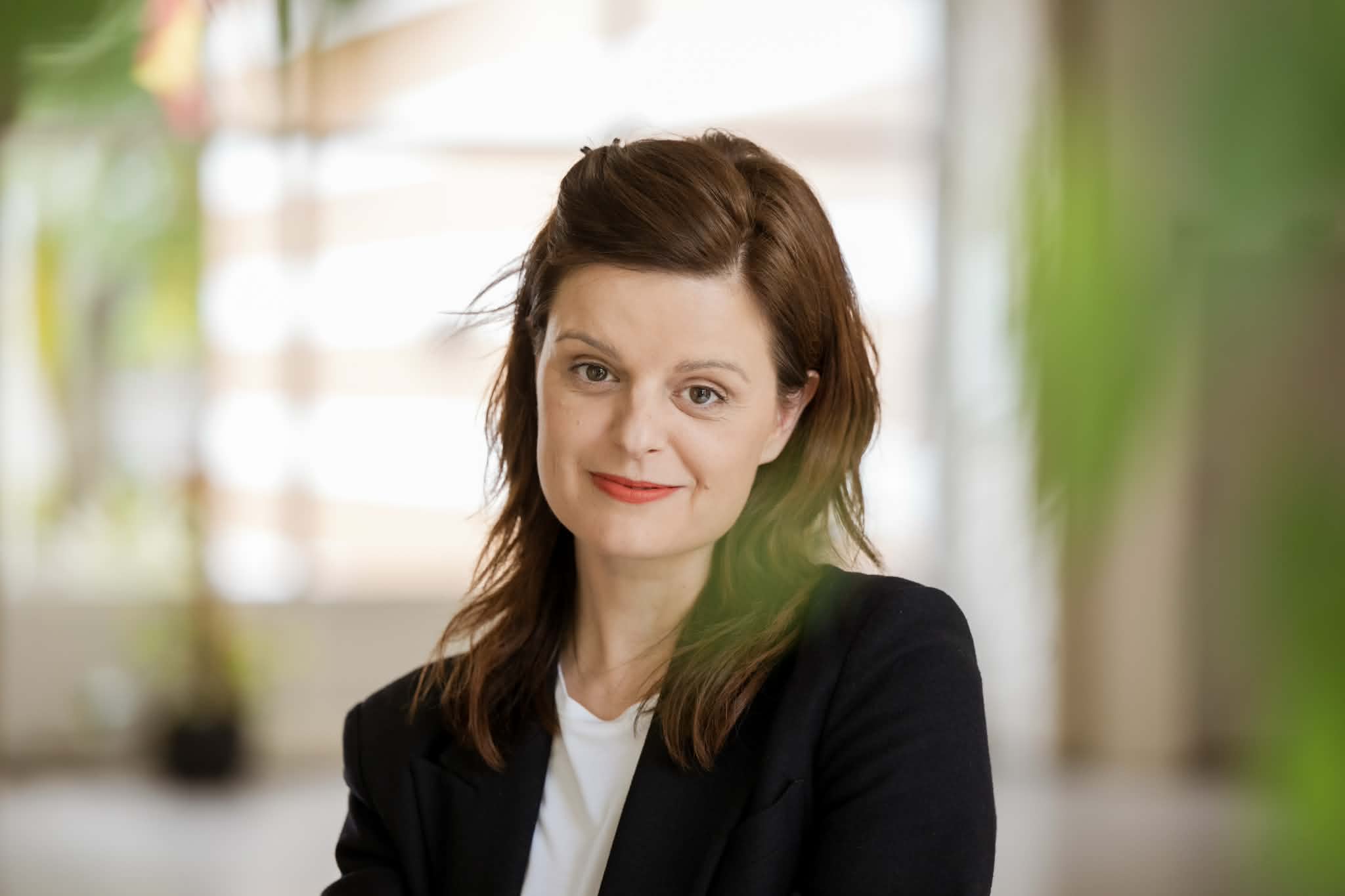 A portrait of Agnė Liucilė Grickevičė. A woman against a bright, softly blurred room background with botanical accents. She wears an unbuttoned black blazer over a white T-shirt and smiles gently, looking directly into the camera. Her medium-length, dark brown hair falls naturally over her shoulders, with the top section pinned back. She wears a soft red lipstick.